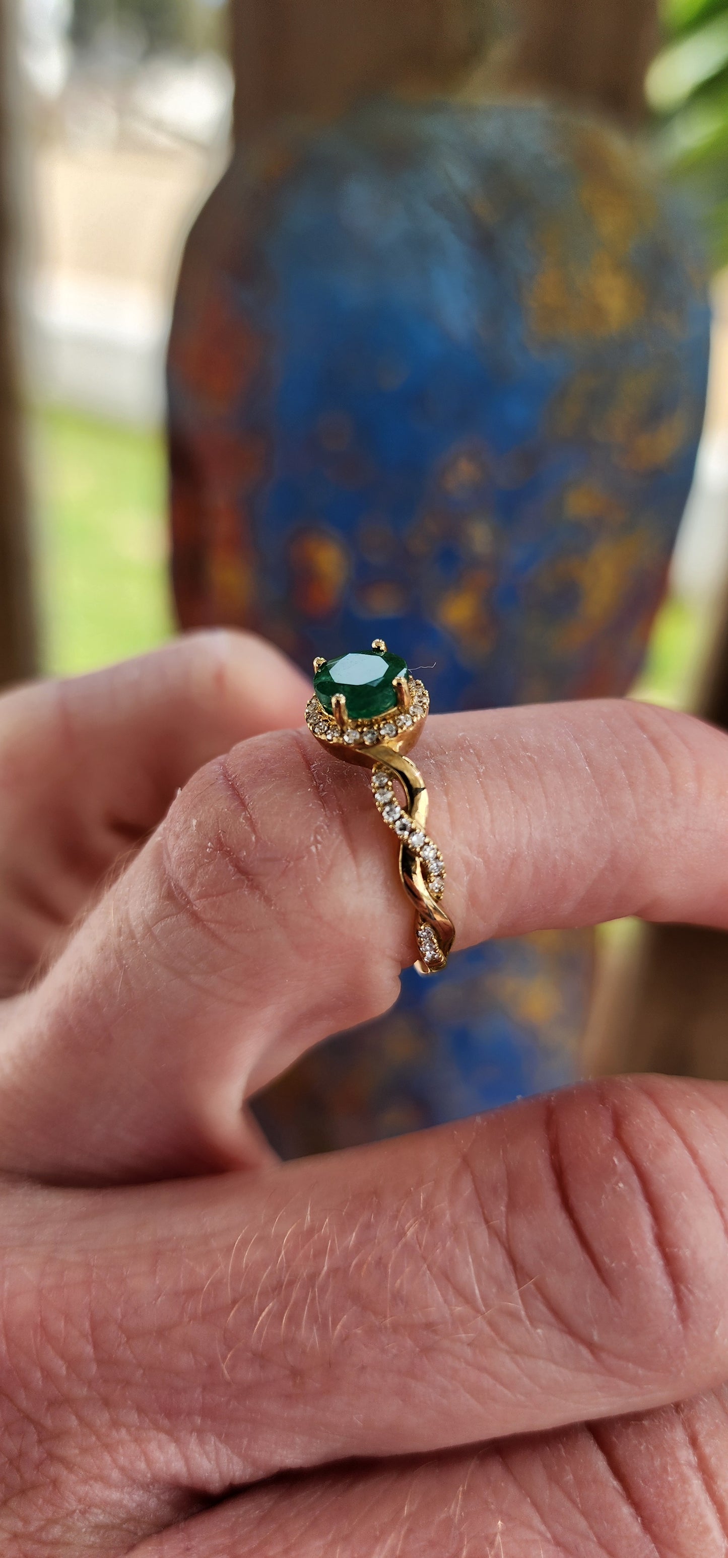 Exquisite 9ct Yellow Gold Crossover/Braided halo Ring, which boasts a mesmerizing prong set Round cut Emerald as its focal point,framed in a halo of small Brilliant Round cut Diamonds.
