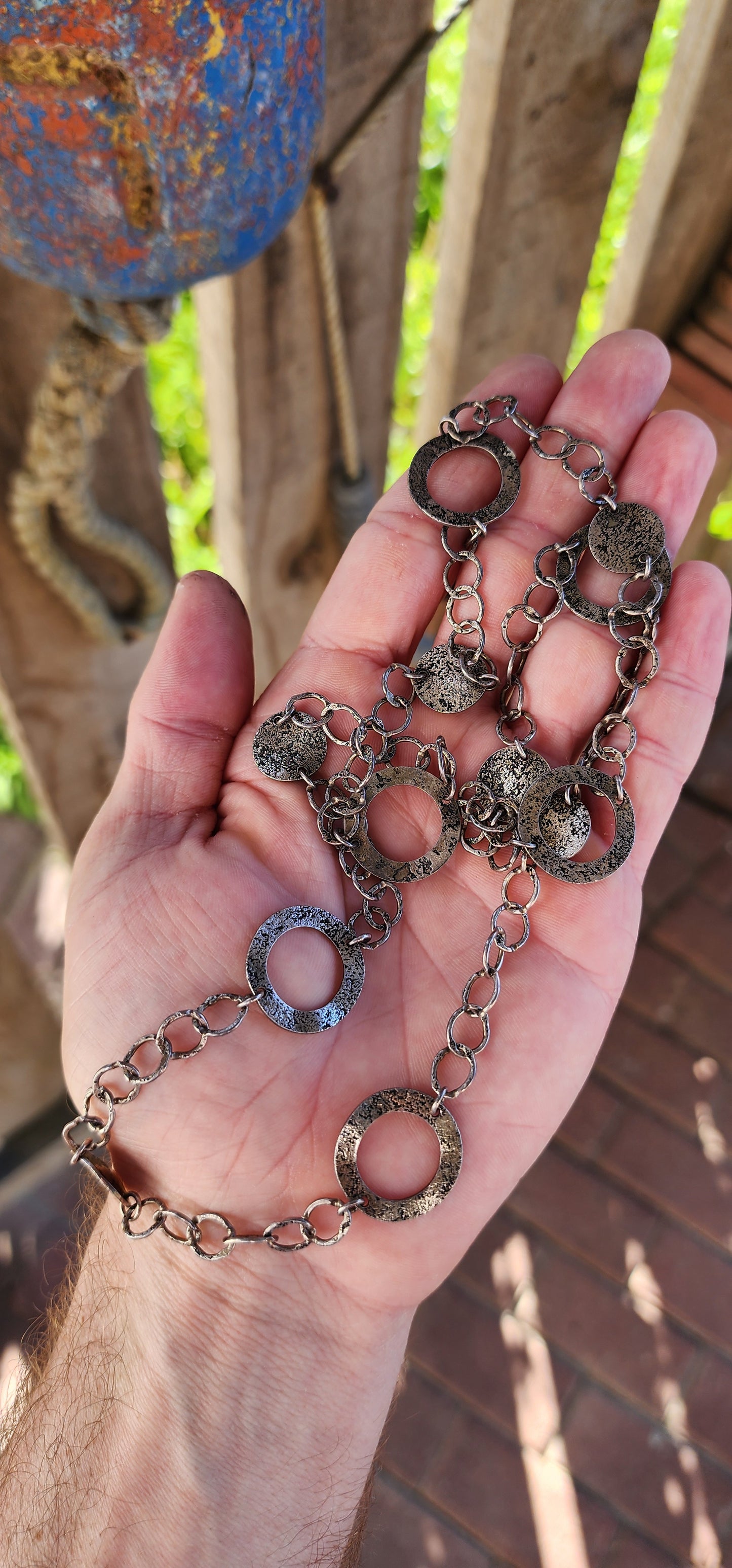 This necklace is a bold, free-spirited statement piece that dances between modernist minimalism and raw, textural brutality. Crafted from sterling silver, it showcases a mesmerizing interplay of hammered, textured, and patinated surfaces.