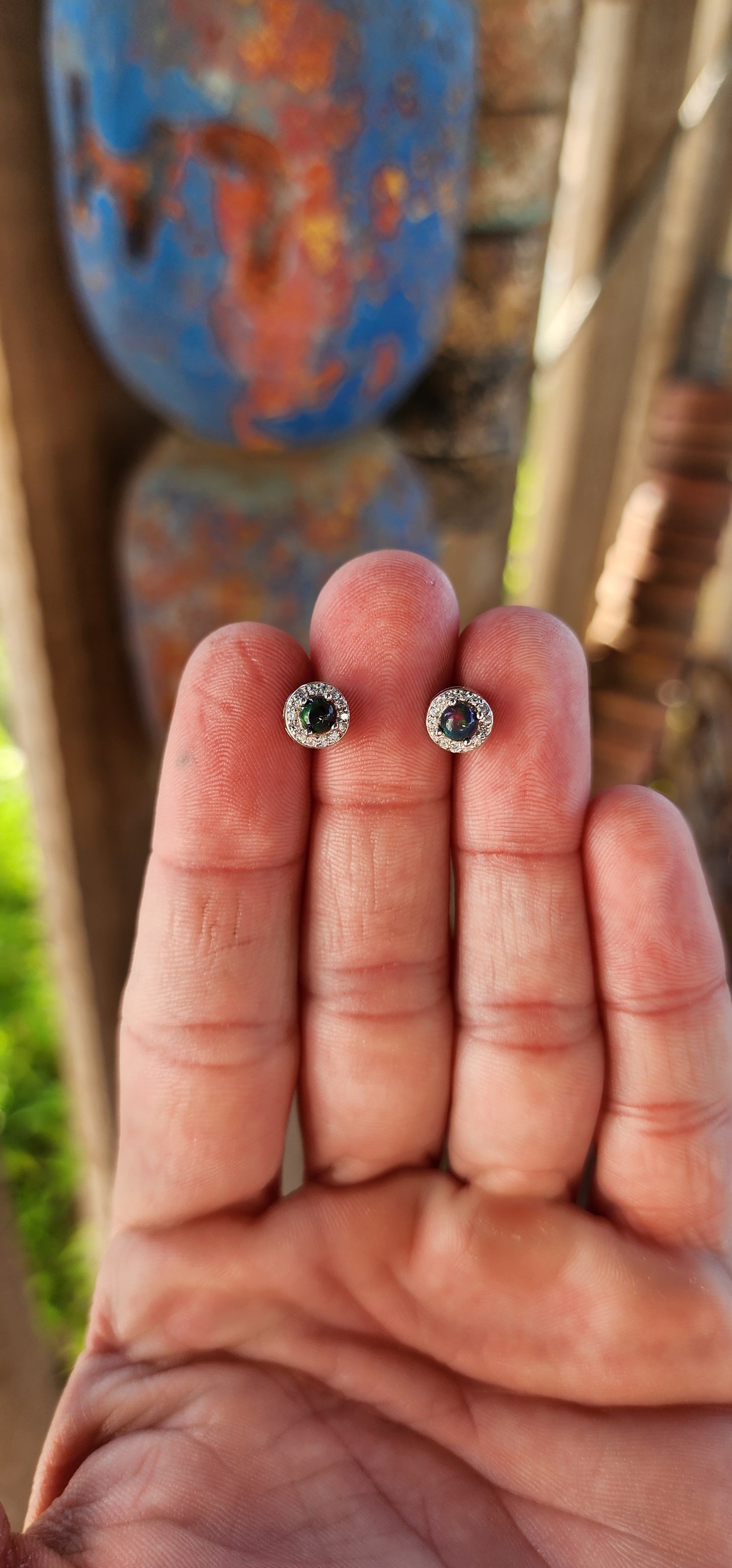 These exquisite Ethiopian black opal halo stud earrings exude classical elegance and refined beauty. Fashioned from sterling silver, each earring showcases a mesmerizing round-cut black opal cabochon, held securely in a delicate prong setting.