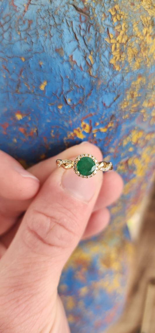 Exquisite 9ct Yellow Gold Crossover/Braided halo Ring, which boasts a mesmerizing prong set Round cut Emerald as its focal point,framed in a halo of small Brilliant Round cut Diamonds.