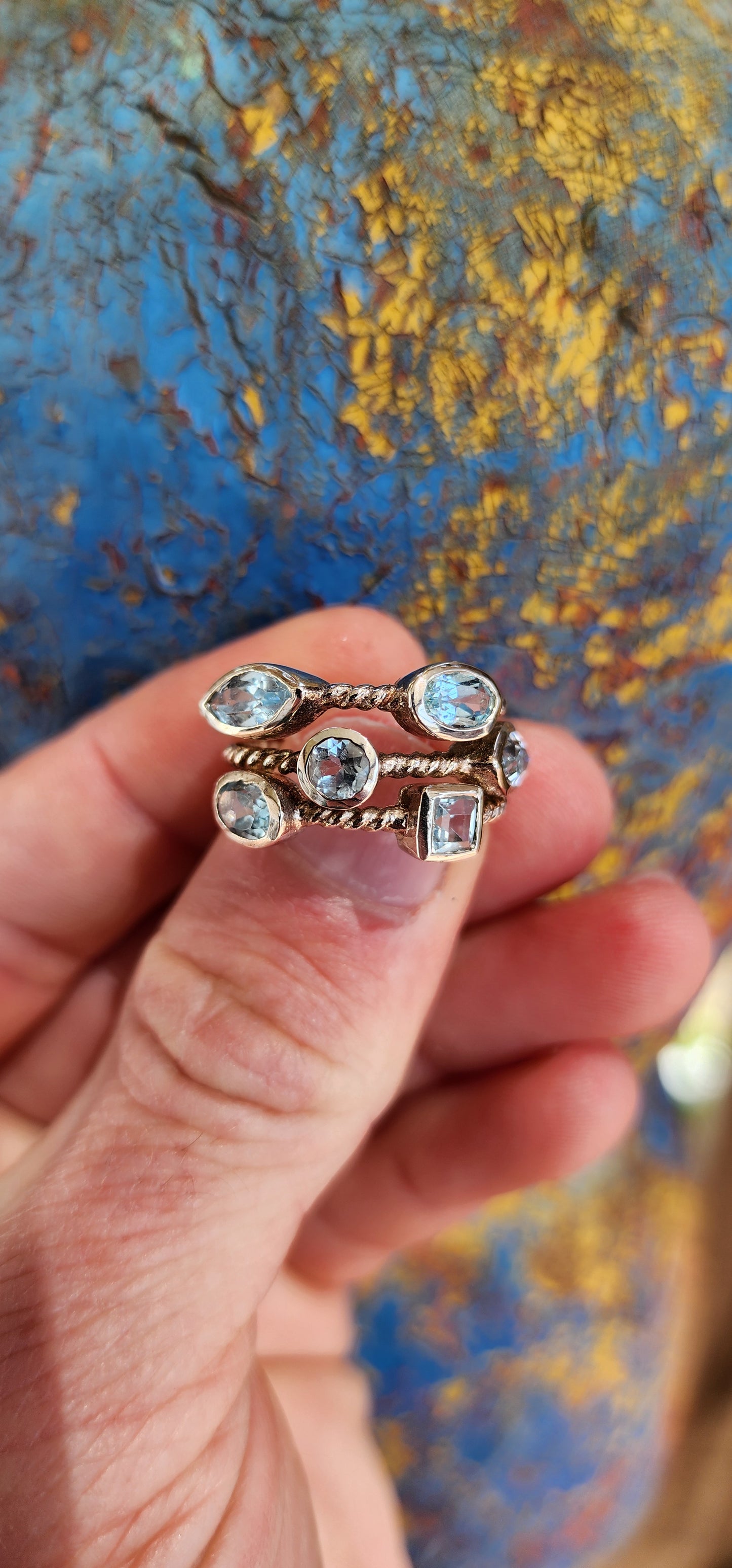 This unique Sterling Silver cluster ring features a Rope pattern on three bands and is adorned with Sky Blue Topaz Gemstones in various shapes and sizes.
The Topaz Gemstones being bezel/tube set.