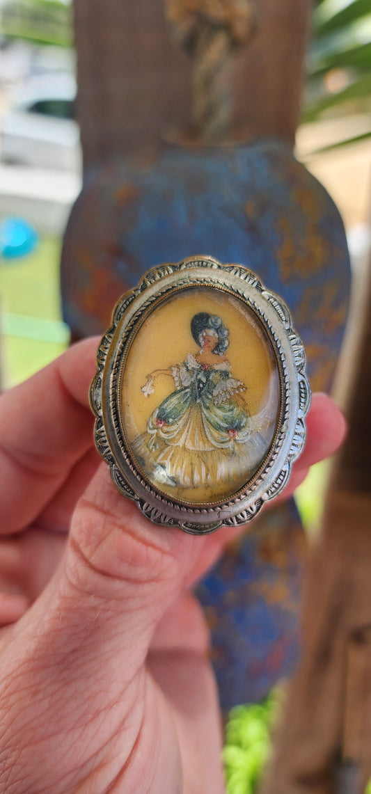 This brooch is an exquisite example of vintage craftsmanship, featuring a signed hand-painted miniature portrait encased in a silver-plated frame with a domed, convex glass cover.