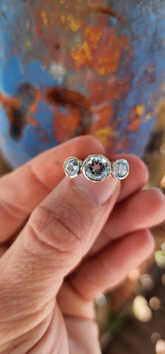 This sterling silver trilogy ring is an elegant embodiment of simplicity and beauty. Its design centers on a radiant round-cut sky blue topaz, tube-set in the middle, which exudes a serene yet mesmerizing glow reminiscent of a clear summer sky.