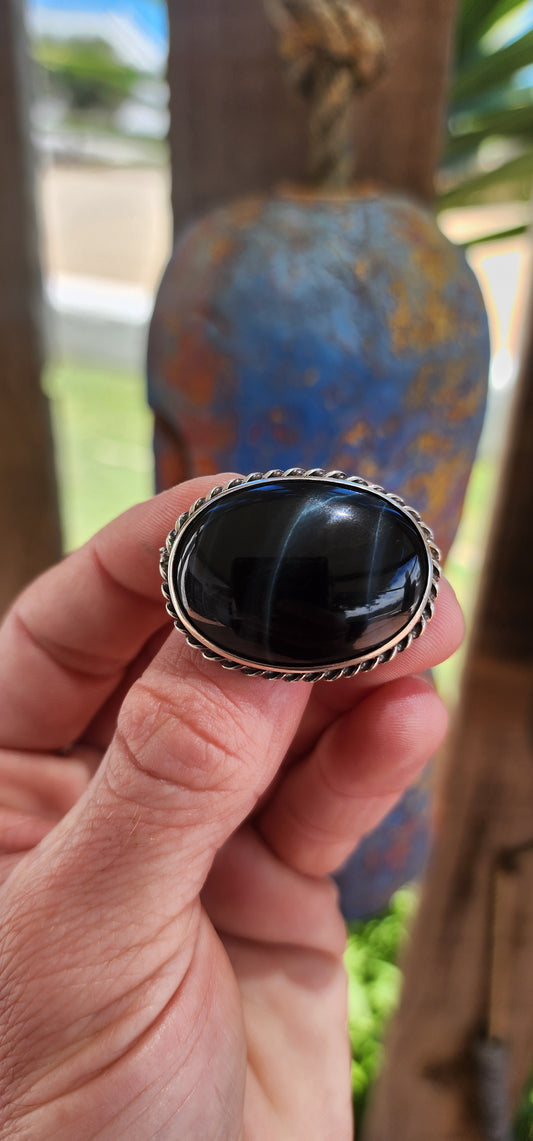 This is a vintage sterling silver oval-shaped brooch featuring a central cabochon of black tiger’s eye. The brooch's sleek and understated design highlights the natural beauty of the stone and the craftsmanship of the silver.