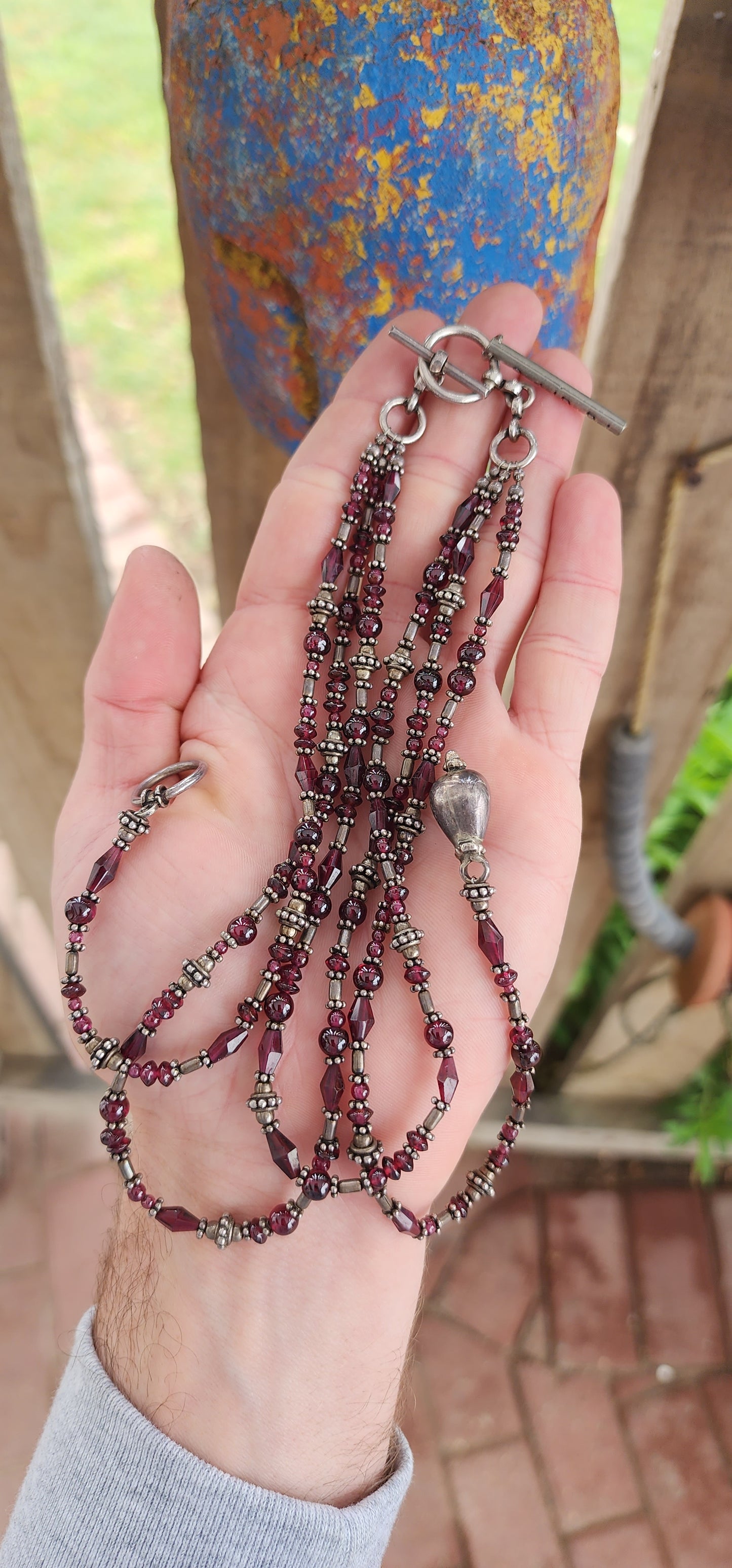 This exquisite artisan-crafted necklace is a stunning three-strand choker that seamlessly blends elegance with versatility. Its intricate design features genuine almandine garnet gemstone beads and sterling silver accents.