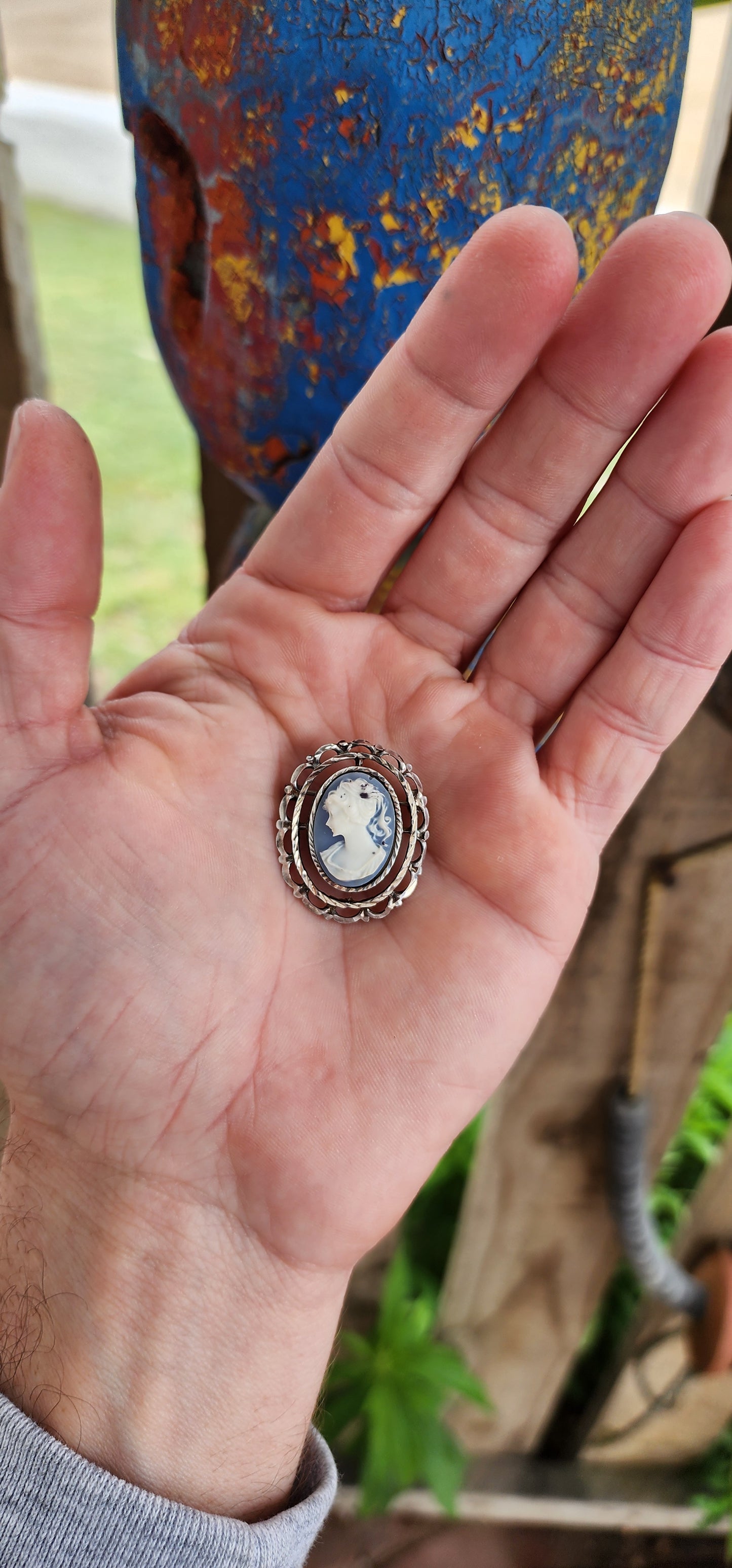 This is a lovely vintage sterling silver brooch featuring a delicate blue cameo crafted from resin.
The centerpiece is an oval blue cameo depicting a classic profile of a lady, likely inspired by Victorian-era motifs.