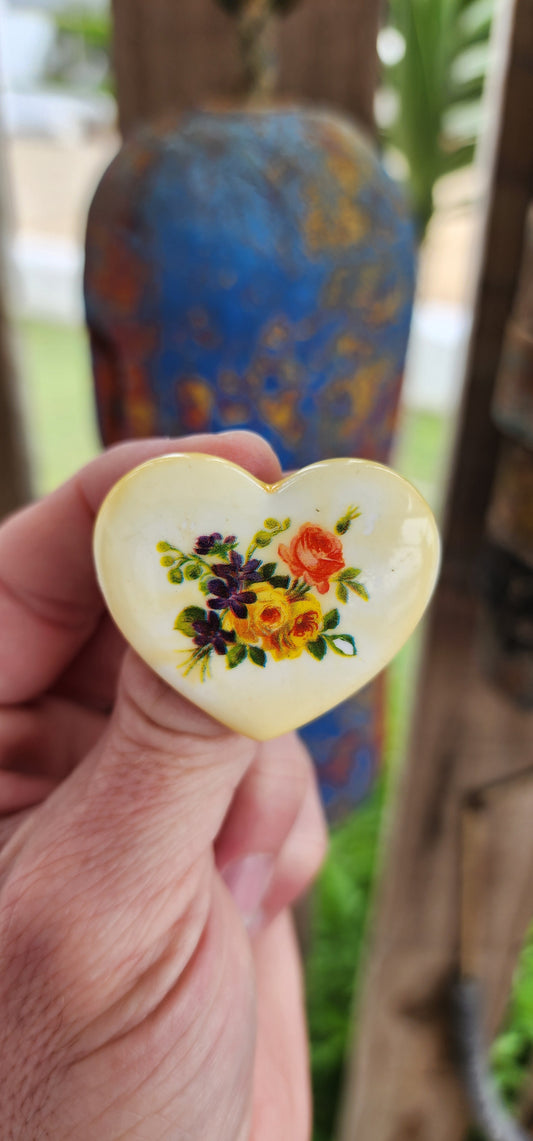 This is a delightful example of vintage Western Germany plastic jewelry, imbued with charm and historical significance. The brooch, shaped like a heart, features a warm ivory background and a meticulously detailed floral motif.