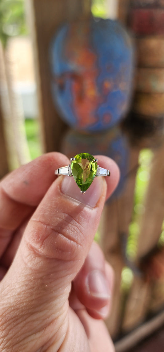 Say hello to this bold beauty — a sterling silver trilogy ring that’s practically glowing with zest and charm!
At its heart lies a mesmerizing, generously sized pear-cut peridot, gleaming in a vivid lime-green hue.