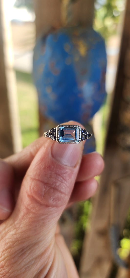 A delicate balance of clarity, color, and craftsmanship, this elegant trilogy-style stacking ring is crafted in sterling silver. At its heart lies a captivating emerald-cut sky blue topaz, its crystalline hue as refreshing as a cloudless morning sky.
