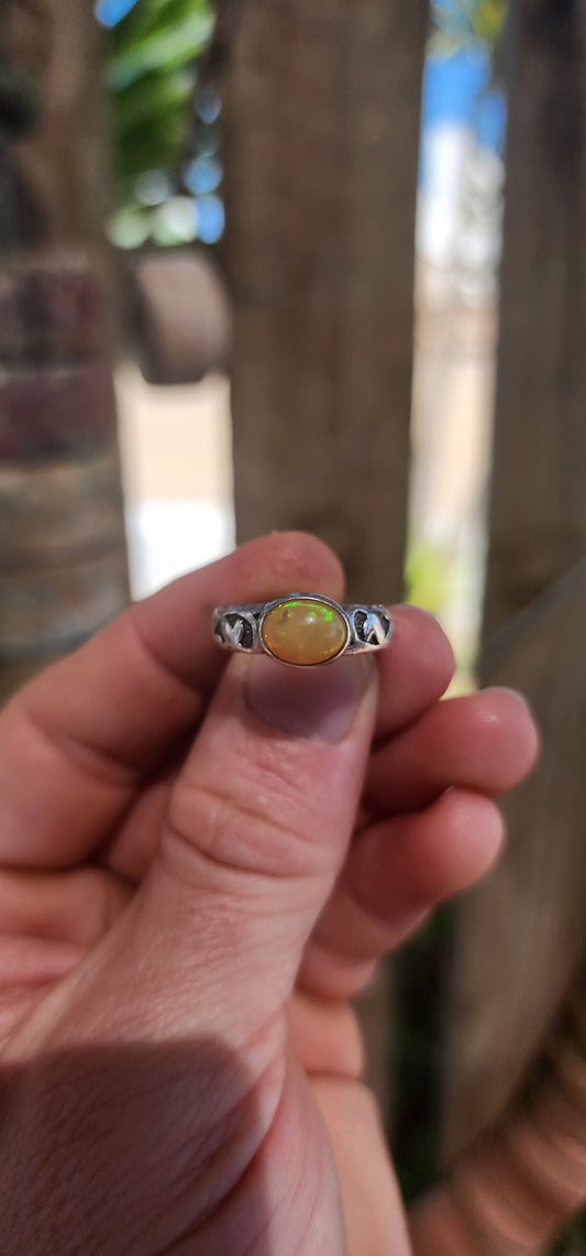 An absolutely mesmerizing handcrafted sterling silver ring featuring a luminous oval-cut Ethiopian fire opal cabochon, radiating with flickers of neon green, orange, and gold as it catches the light.