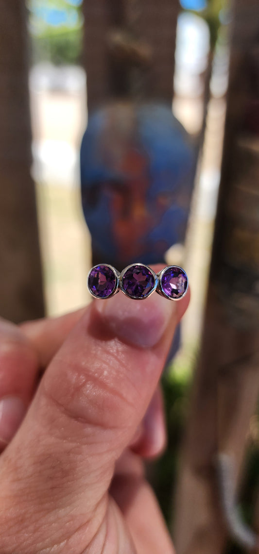 This sterling silver trilogy ring is a dreamy celebration of amethyst magic, set with three round faceted amethyst gemstones that glow in juicy shades of violet, plum and royal purple.