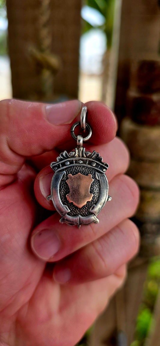 This beautiful antique shield fob pendant is crafted in hallmarked sterling silver, featuring a striking 9ct rose gold shield cartouche at its centre.