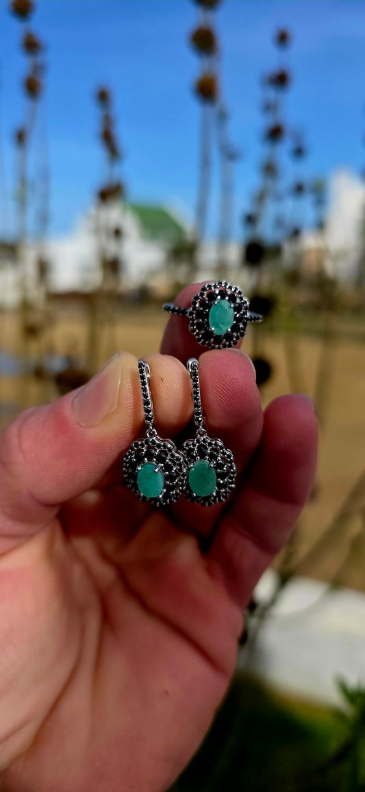 Beautiful matching sterling silver floral-inspired ring and earring set featuring luminous oval-cut emerald gemstones at their centres, each delicately prong set and framed in dramatic double scalloped halos of sparkling black spinel gemstones.