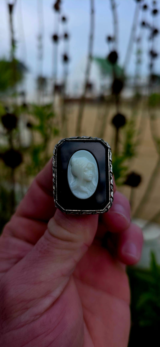 This beautiful vintage brooch features a striking oval milk glass cameo depicting the graceful profile of a young lady, her delicate features sculpted in crisp relief against a dramatic black glass background.