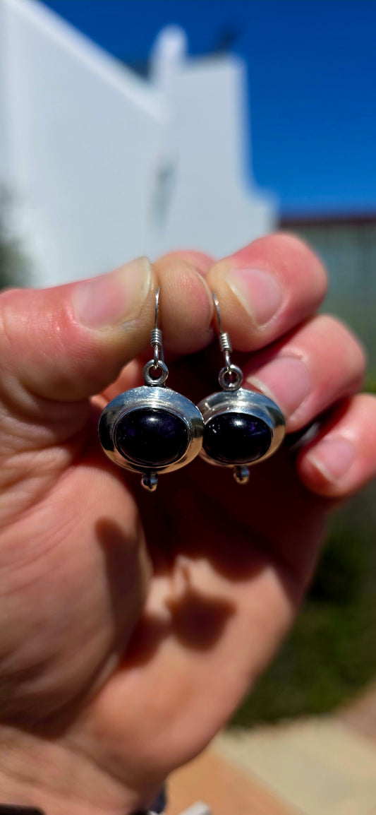 A beautiful pair of artisan-style sterling silver drop earrings featuring deep purple amethyst cabochons, beautifully complemented by sculptural silver settings.